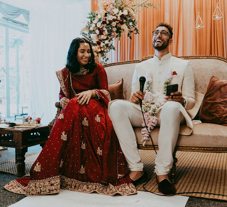 Bride wears red and gold embellished bridal separates as she sits beside her groom in white sherwani with red pocket square 