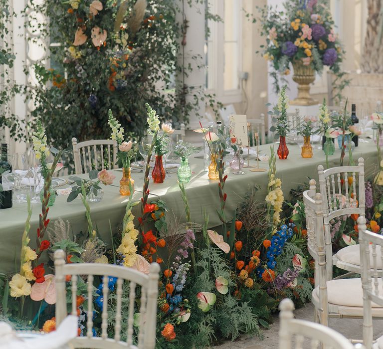 Brightly coloured floral arrangements with green foliage in front of head table with pastel green tablecloth and floral installation to back