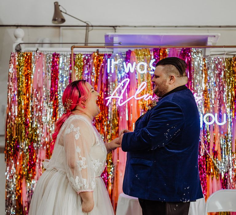 Groom in midnight blue velvet blazer and bride in corset top leaf detailed lace overlay wedding dress laughing at the alter standing in front of metallic streamer backdrop 