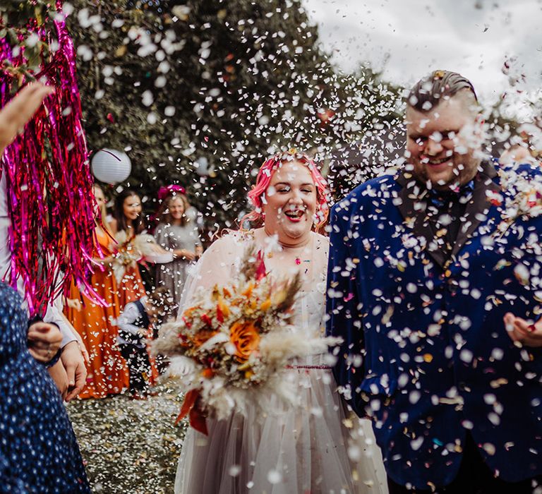 Bride in leaf detailed lace overlay wedding dress with autumnal bouquet with pampas grass and orange ribbon holding hands with groom in midnight blue velvet suit with autumnal boutonniere doing confetti walk