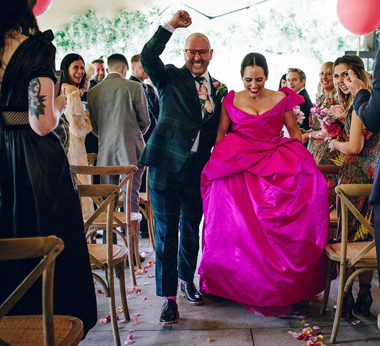 Groom doing fist pump in dark forest green tartan suit with patterned tie, white pocket square and pink and green boutonniere and bride in off the shoulder front ruching pink wedding dress walking down the aisle at Middleton Lodge wedding venue 