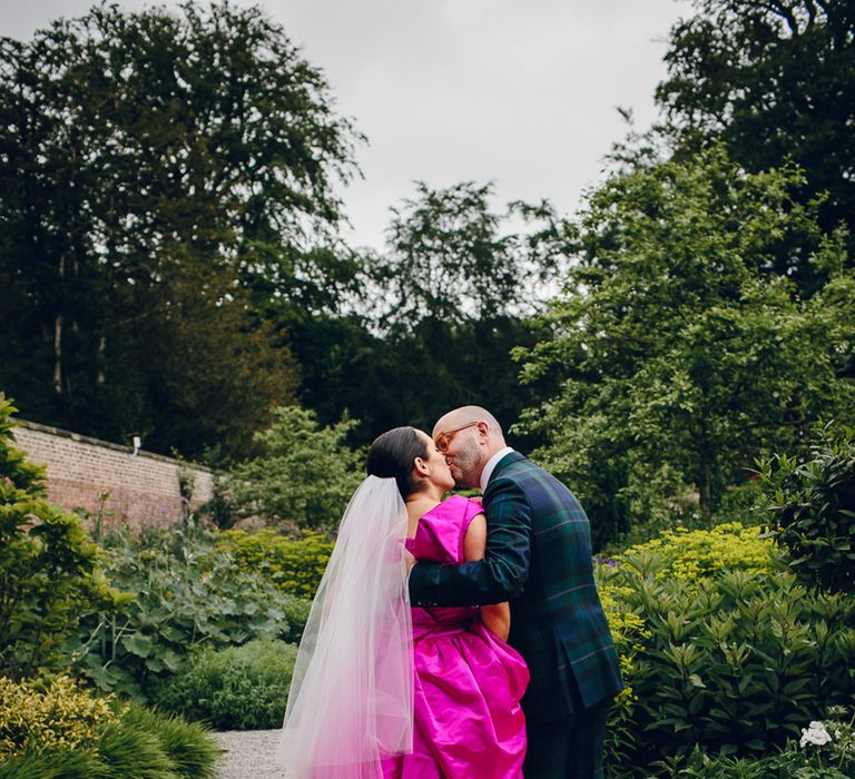 Groom in dark forest green tartan suit with patterned tie, white pocket square and pink and green boutonniere kissing bride in off the shoulder front ruching pink wedding dress standing in the gardens of Middleton Lodge 