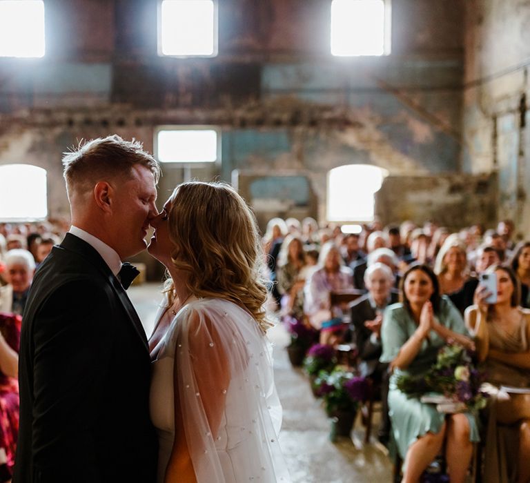 Bride in pearl encrusted wedding cape leans in to kiss her groom during wedding ceremony