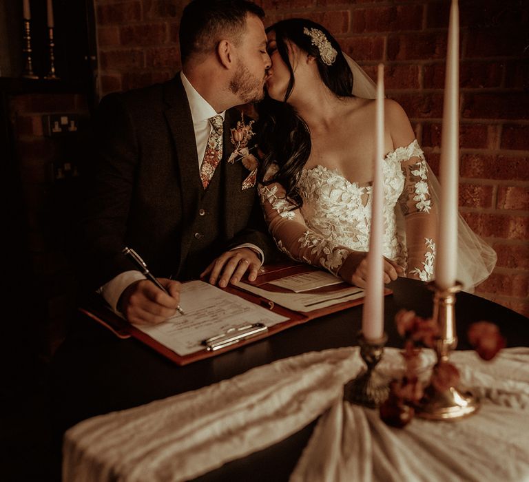 Groom in flower patterned tie and pocket square and dried flower boutonniere signing the register and kissing bride in off the shoulder bodycon wedding dress with long, sheer sleeves and lace detailing and large silver and diamond hair accessory 