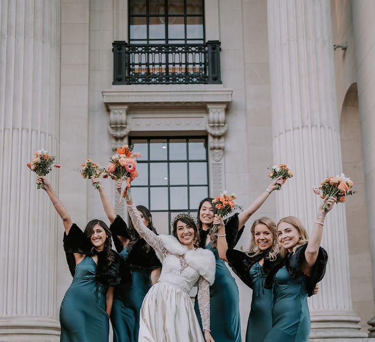 Bride in taffeta material princess wedding dress with long lace sleeves  and cropped faux fur bridal cover up holding up bridal bouquet with bridesmaids in deep green satin bridesmaid dresses with faux black coverups and black open toe heels 