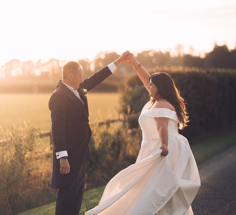 Groom in a navy morning suit with a white rose buttonhole and tie spinning the bride around as they dance together during sunset with the bride in an off the shoulder wedding dress and emerald and gold bracelet 