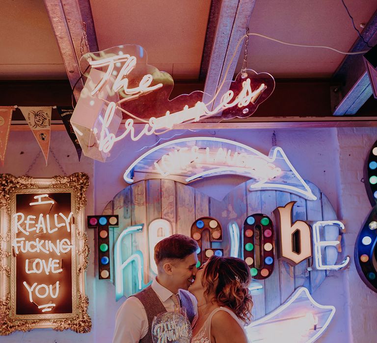 Groom in grey three piece wedding suit with yellow suit and yellow pocket square and bride in sleeveless plunge lace wedding dress and cathedral length embroidered veil kissing in front of numerous neon signs and LED signs at Gods Own Junkyard 