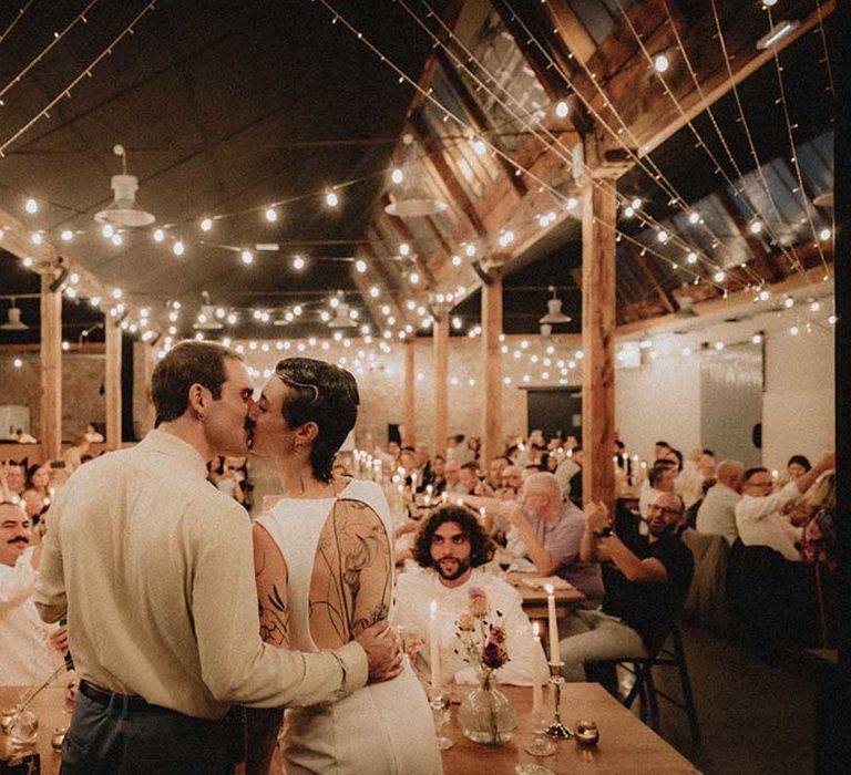 Lisa & Gabriel kiss in front of wedding guests during reception in industrial setting with festoon lighting 