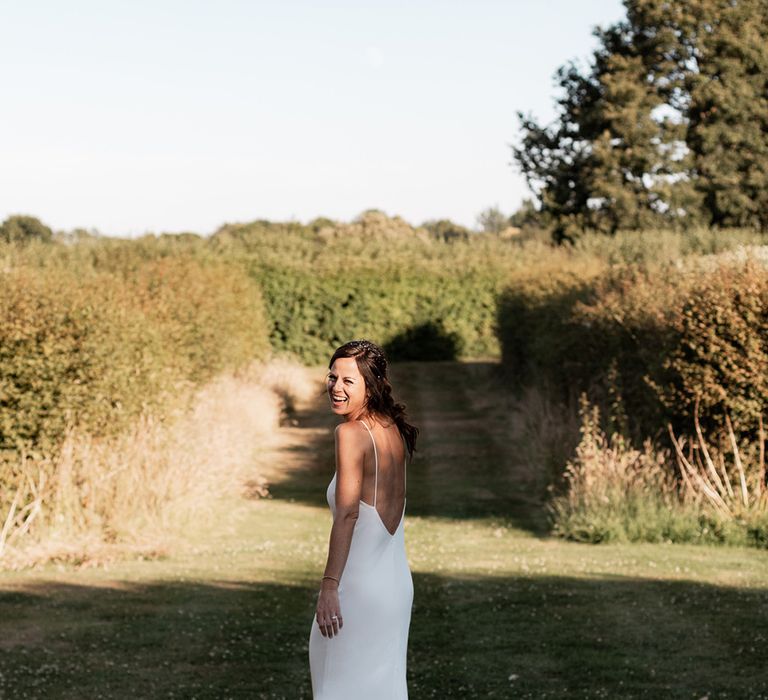 Bride wears simple silk low back wedding dress and her brown hair in low up-do with loose curls 