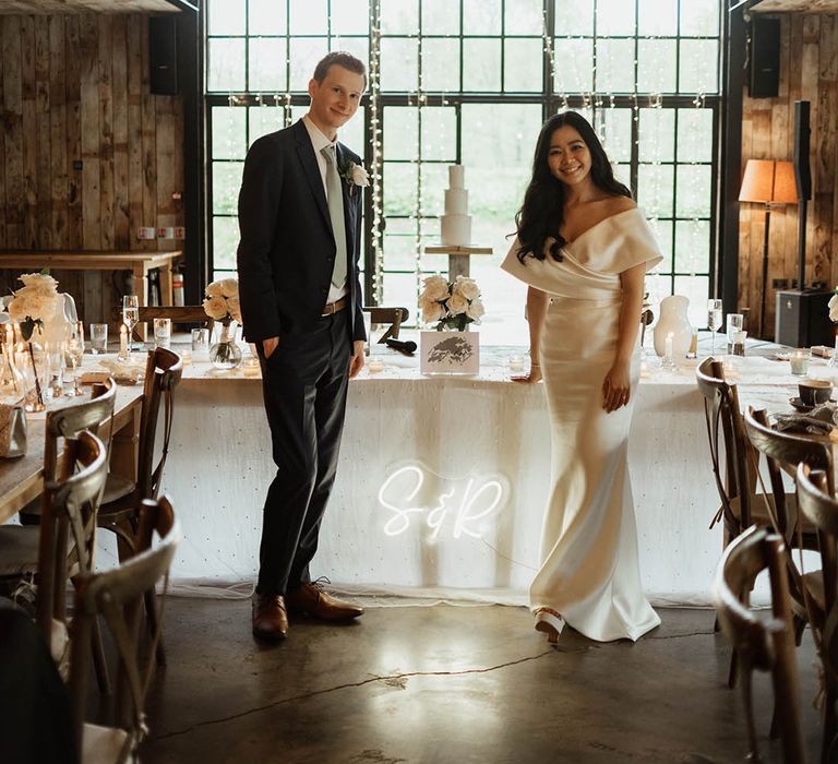 The bride and groom smile together next to their personalised neon sign and their white wedding cake decorated with pearls 