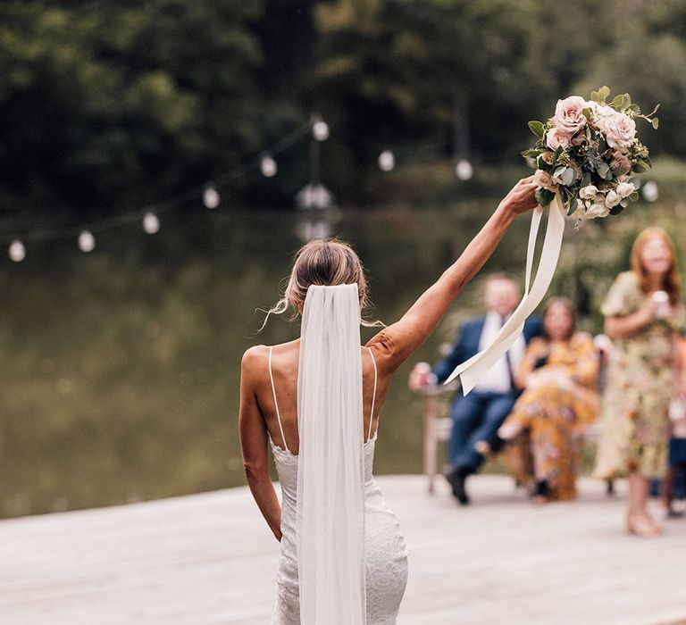 The bride in a fitted lace wedding dress with a veil lifts her arm with a pink garden rose bouquet with white carnation 