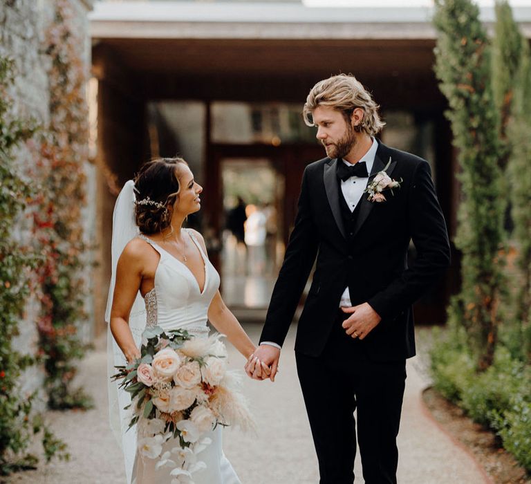 The groom in a black tuxedo walks holding hands with the bride in an elegant satin finish mermaid wedding dress 