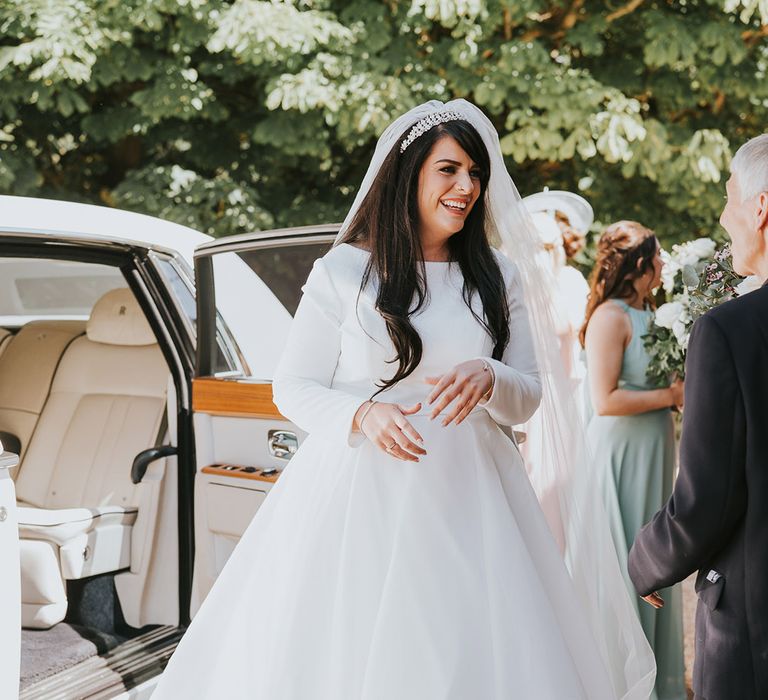 Bride with long brown hair gets out of the white wedding car transport in a high neck long sleeve wedding dress with tiara and veil accessory 