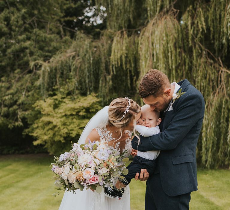 The bride in a Dando London wedding dress kissing the baby boy held by the groom in a navy suit 