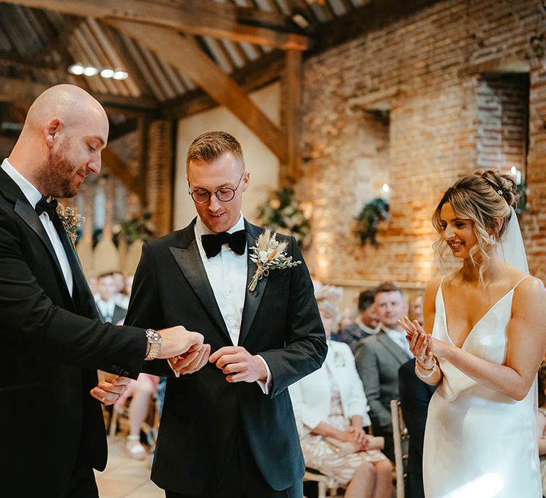 Best man in black tuxedo passes the rings to the groom also wearing a black tuxedo and glasses for the civil ceremony 