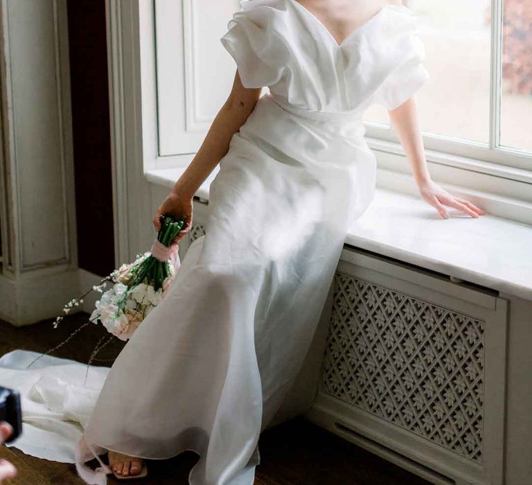 Bride wearing layered, sculpted off the shoulder wedding dress and open toe wedding heels holding spring inspired bridal bouquet 