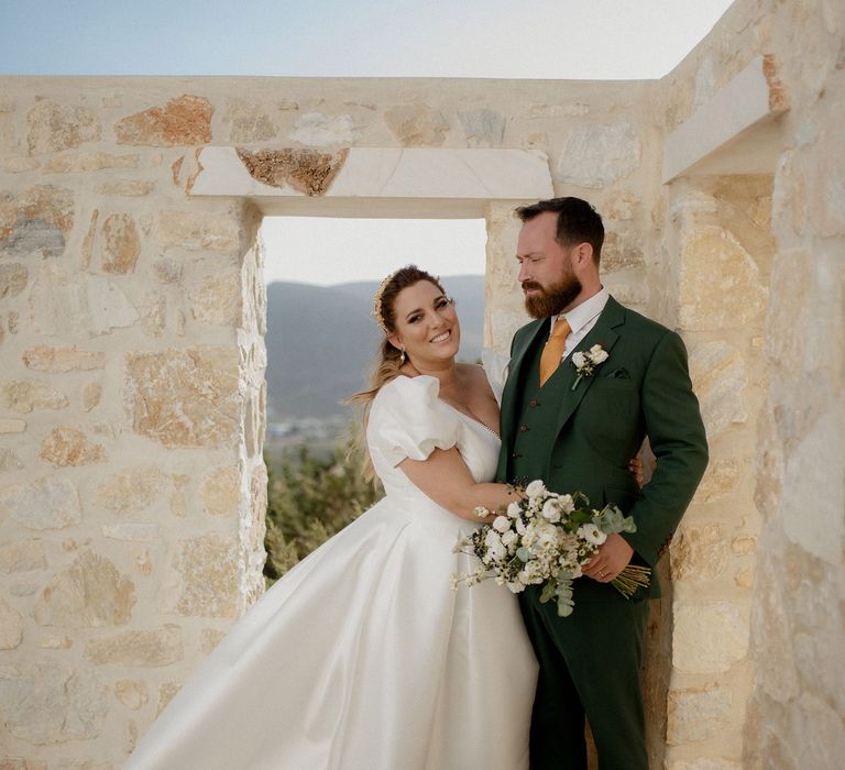 Bride in white satin princess dress featuring puff-sleeves poses with groom in dark green wedding suit at private villa wedding in Greece