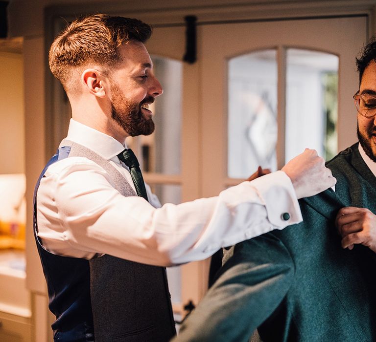 Groomsman helps the groom into his dark coloured suit jacket with dark brown tie 