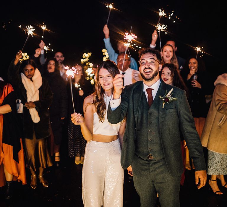 Bride in sequin trousers and top bridal separates with the groom waving sparklers for sparkler send off 