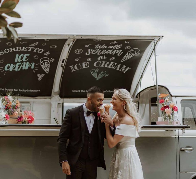 Bride in off the shoulder lace wedding dress standing with groom in classic three piece grooms tuxedo with colourful spring boutonniere eating ice-cream standing by wedding food truck 