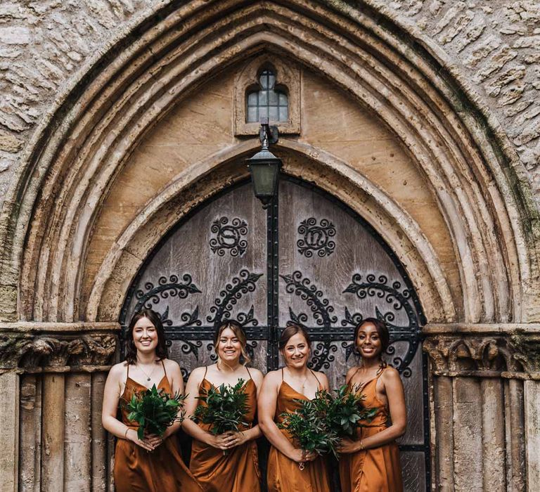 Bridesmaids standing outside of the church wearing strappy sleeveless front slit burnt orange bridesmaid dresses holding foliage bouquets 