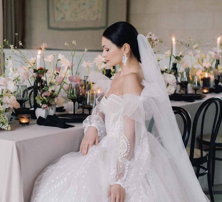 Bride in off the shoulder white and pink floral patterned bridal gown with black bow and front slit sitting at black white and pink wedding tablescape with light pink wedding tablecloth, white poppies, light pink Persian buttercups, baby's-breath and foliage centrepieces, black artistic candlestick holders, white tapered candles, black crockery and monochrome wedding stationery 