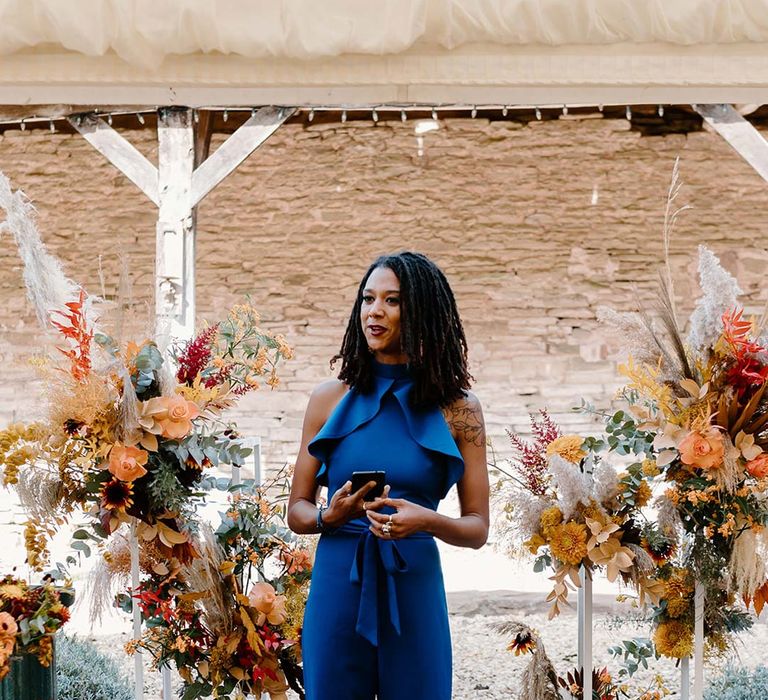 Wedding guest leading the wedding ceremony wearing cobalt blue jumpsuit standing in front of wedding alter flower arrangements with sunflowers, garden roses, pampas grass, eucalyptus, yellow tall kangaroo paws, orange osmanthus flowers, foliage and dried flowers 