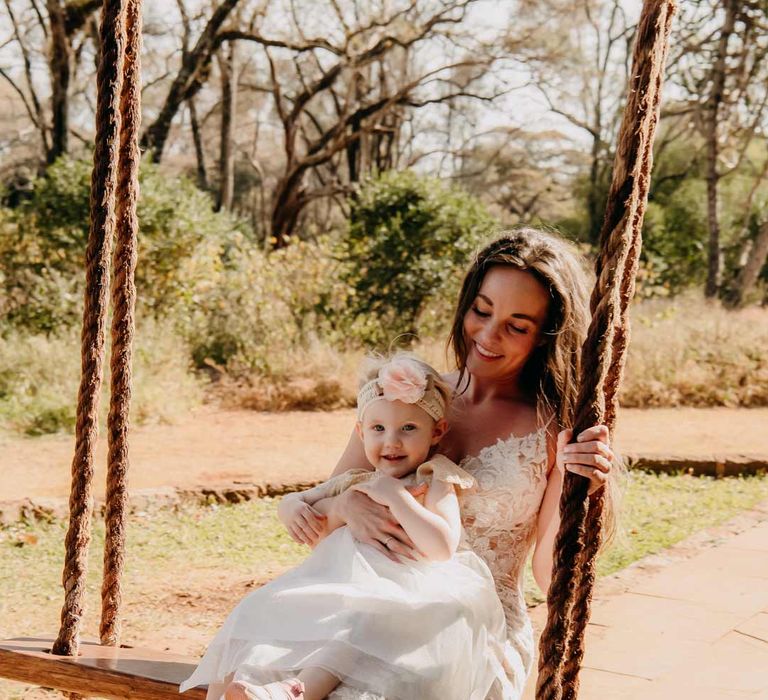 Bride in lace sleeveless wedding dress with puddle train holding child in off-white dress and sequin headband with pink carnation swinging on a swing at Giraffe Manor wedding venue