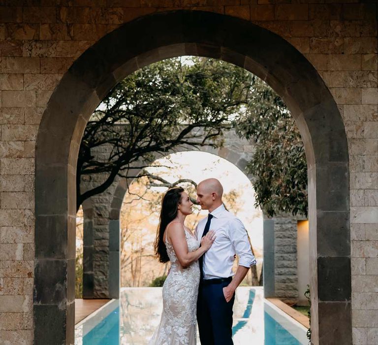 Groom in white shirt, black tie and black suit trousers kissing bride in lace sleeveless wedding dress with puddle train by the swimming pool at Giraffe Manor wedding venue