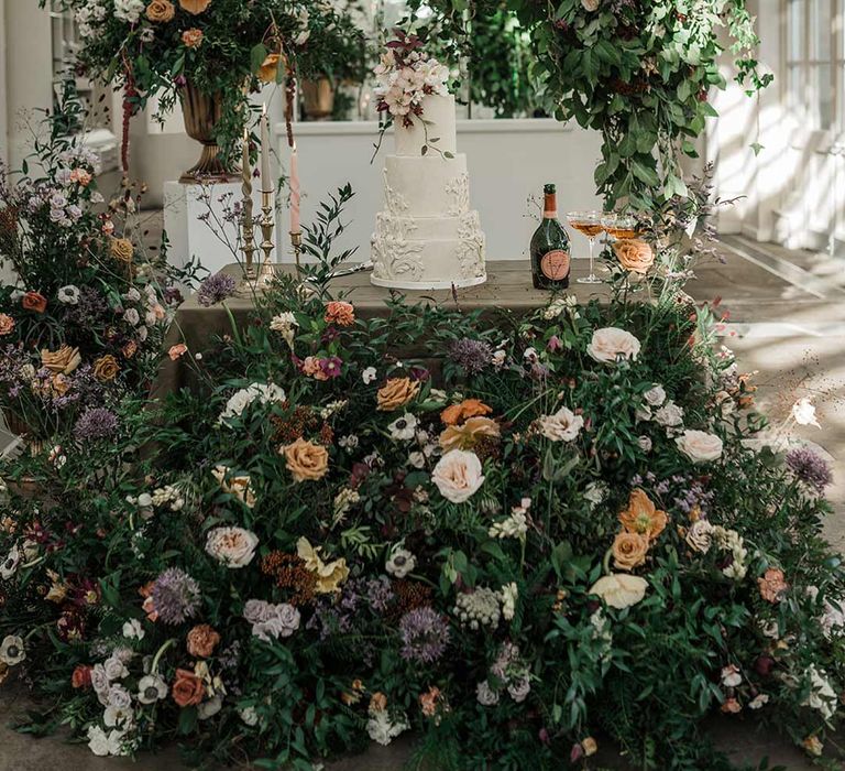 Four tier white wedding cake with intricate icing details and dried flower wedding cake topper on wooden table with twisted tapered candles in neutral shades in gold candlesticks surrounded by wildflower and foliage wedding flower arrangements