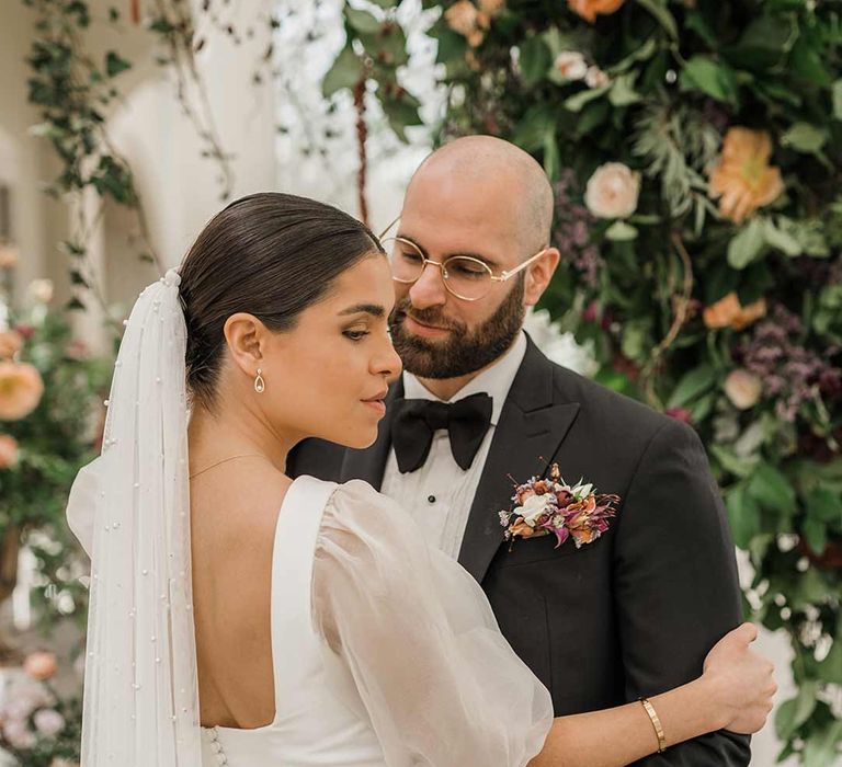 Bride in short sheer puff sleeve wedding dress with square neck and wrist loop with sheer pearl embellished bridal veil with button detailing dancing with groom in classic black tuxedo with bowtie and wildflower boutonniere