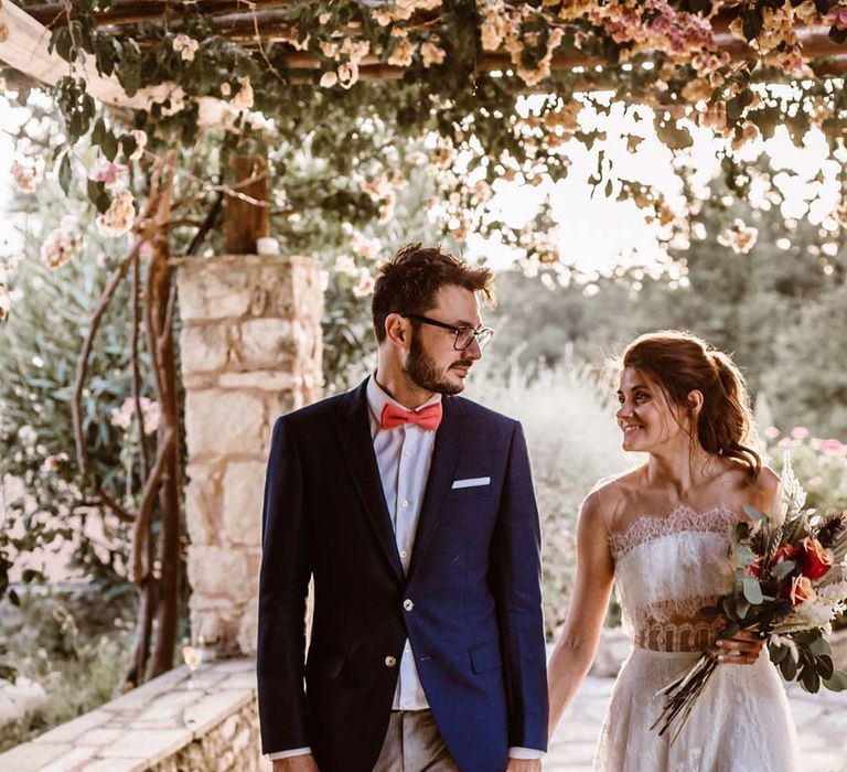 groom in grey chinos navy blazer and orange bow tie holding hands with his bride in a strapless lace wedding dress at greece destination wedding