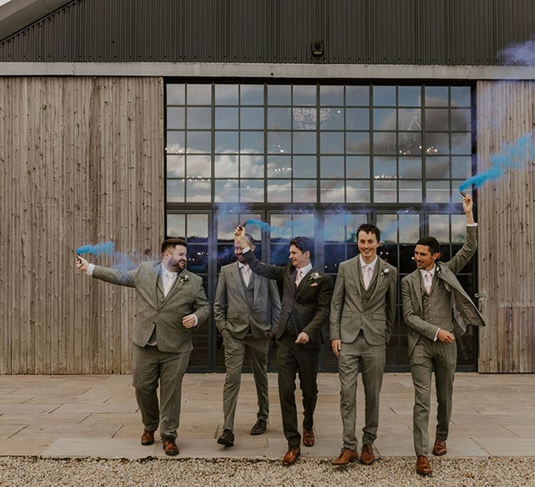 Groomsmen in light grey suits walking with groom waving blue smoke flare 