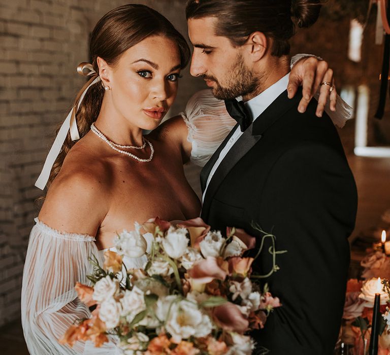 Bride carrying blush pink wedding bouquet with hair ribbon and pearl jewellery posing with the groom in black tuxedo 