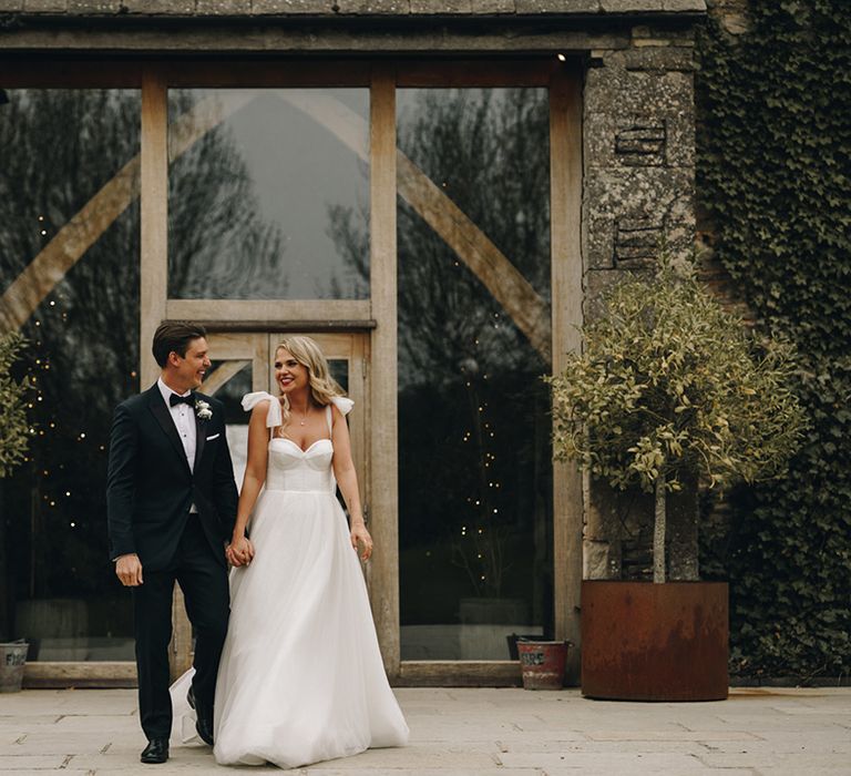 Groom in black tie wedding outfit walking with the bride in corset style wedding dress at Stone Barn