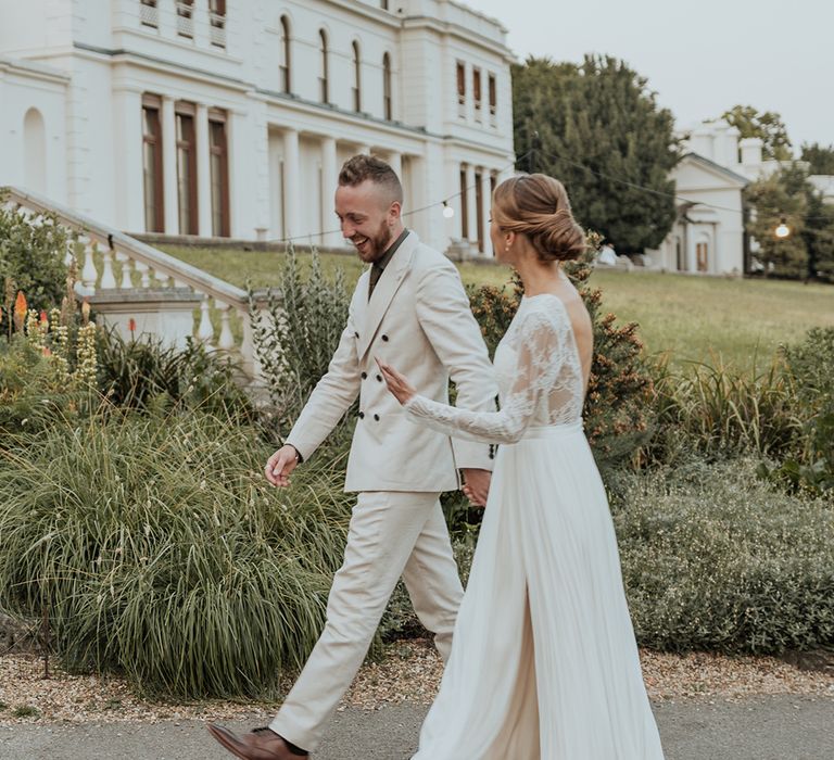 bride in a Catherine Dean floaty skirt wedding dress with front slit and lace long sleeves holding hands with her groom in a beige suit 