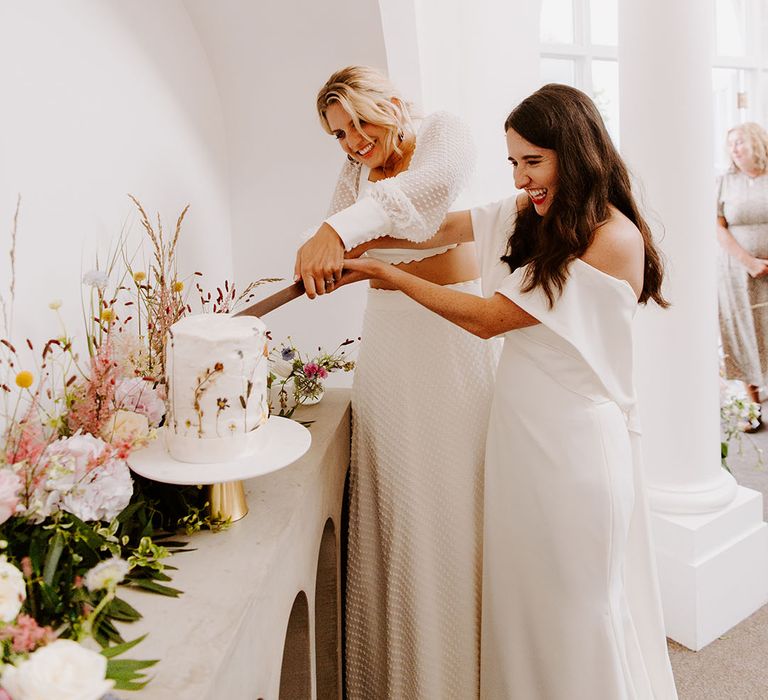 The two brides cut their single tier white iced rustic wedding cake with pressed wildflowers 