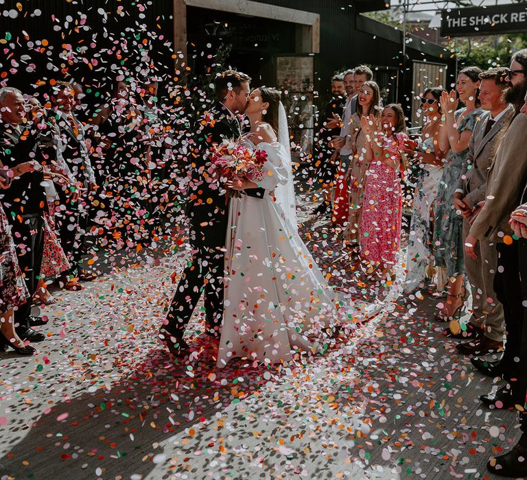 The bride and groom share a kiss as their guests throw the confetti over the couple to celebrate their wedding 