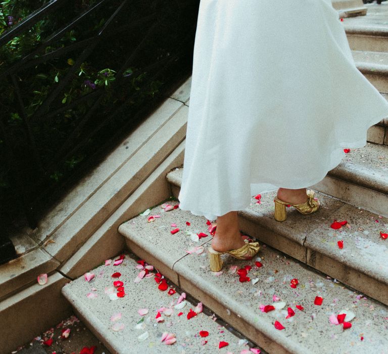 Bride walks up confetti and rose petal covered stairs in slip-on golden bow heels