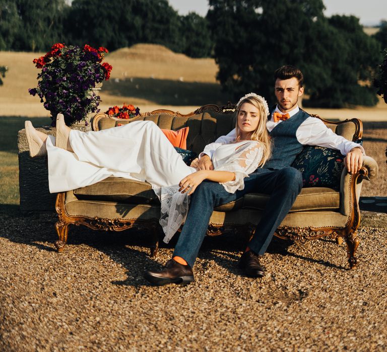 Groom in denim style trouser poses with bride in boho wedding cover and wedges on a plush sofa outside with a purple and red flowers