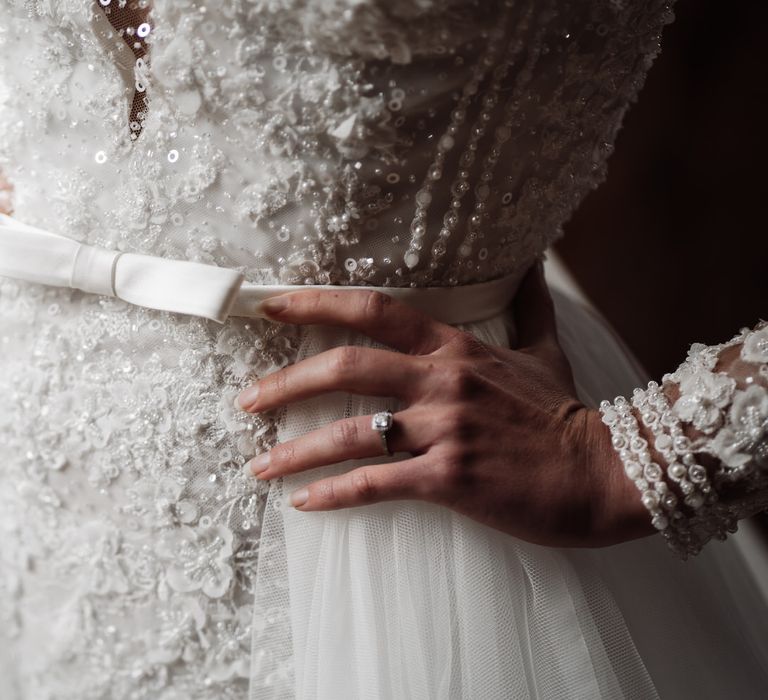 The bride's dress has intricate lace and bead detailing with a bow around the waist.