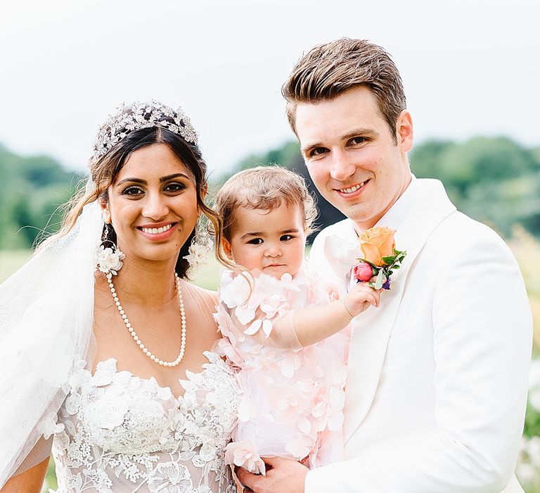 Family portrait with the bride in an appliqué wedding dress and the groom in a white tuxedo jacket holding their flower girl daughter 