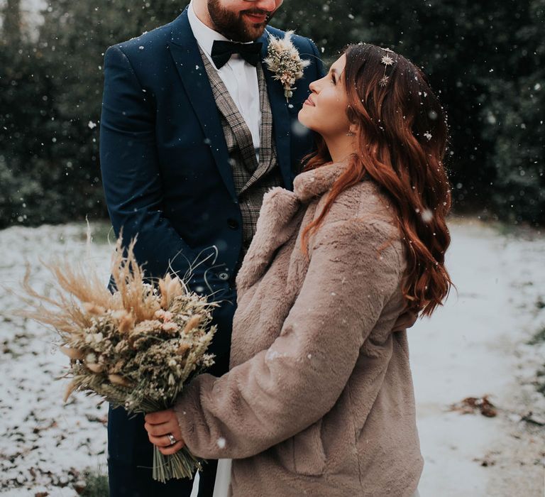 Bride in fur coat holding dried flower bouquet smiles up at groom in navy suit and bowtie at snowy wedding