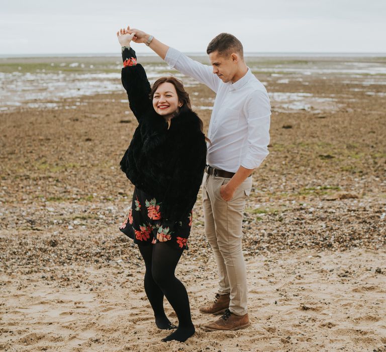 Groom-to-be in a white shirt and chino's twirling his bride-to-be around on the beach in a floral dress, tights and black coat 