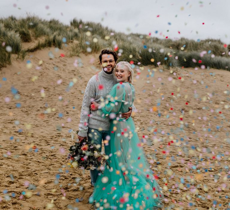 Colourful confetti cannon moment for a beach elopement with bride in a sheer wedding dress and the groom in jumper and jeans 