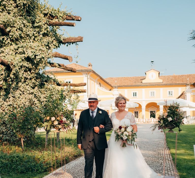 The bride smiling arm in arm with her father as he walks her down the aisle at her outdoor Italian ceremony