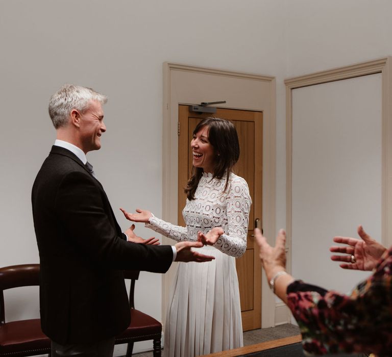 Bride in long sleeved lace top Self Portrait dress, groom in brown woollen suit and registrar all gesture happily at Bristol Registry Office wedding