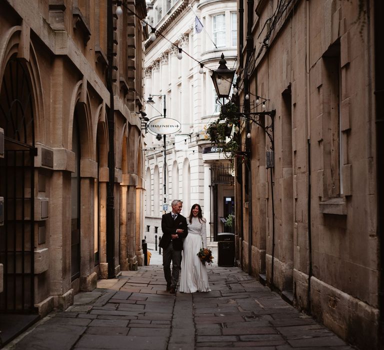 Bride in long sleeved lace top Self Portrait wedding dress holds hands with groom in brown woollen blazer walking through streets of Bristol