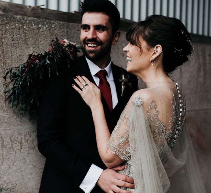 A bride laughs with her new husband as he looks to the side. She wears a homemade wedding dress with a cape.