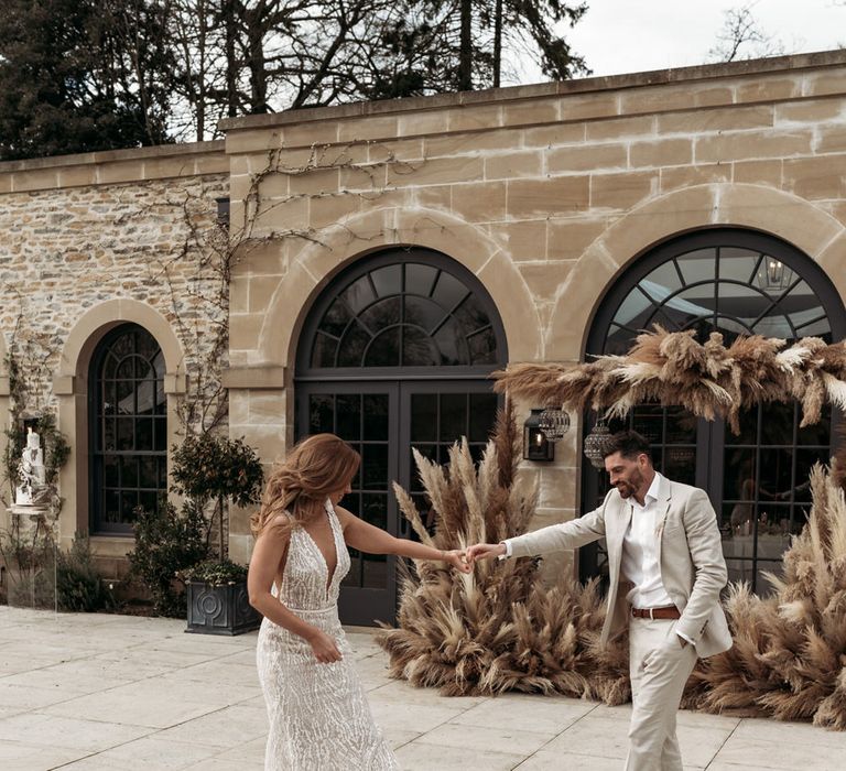 Groom in a beige suit and bride in an embellished wedding dress dancing in front of a pampas grass installation